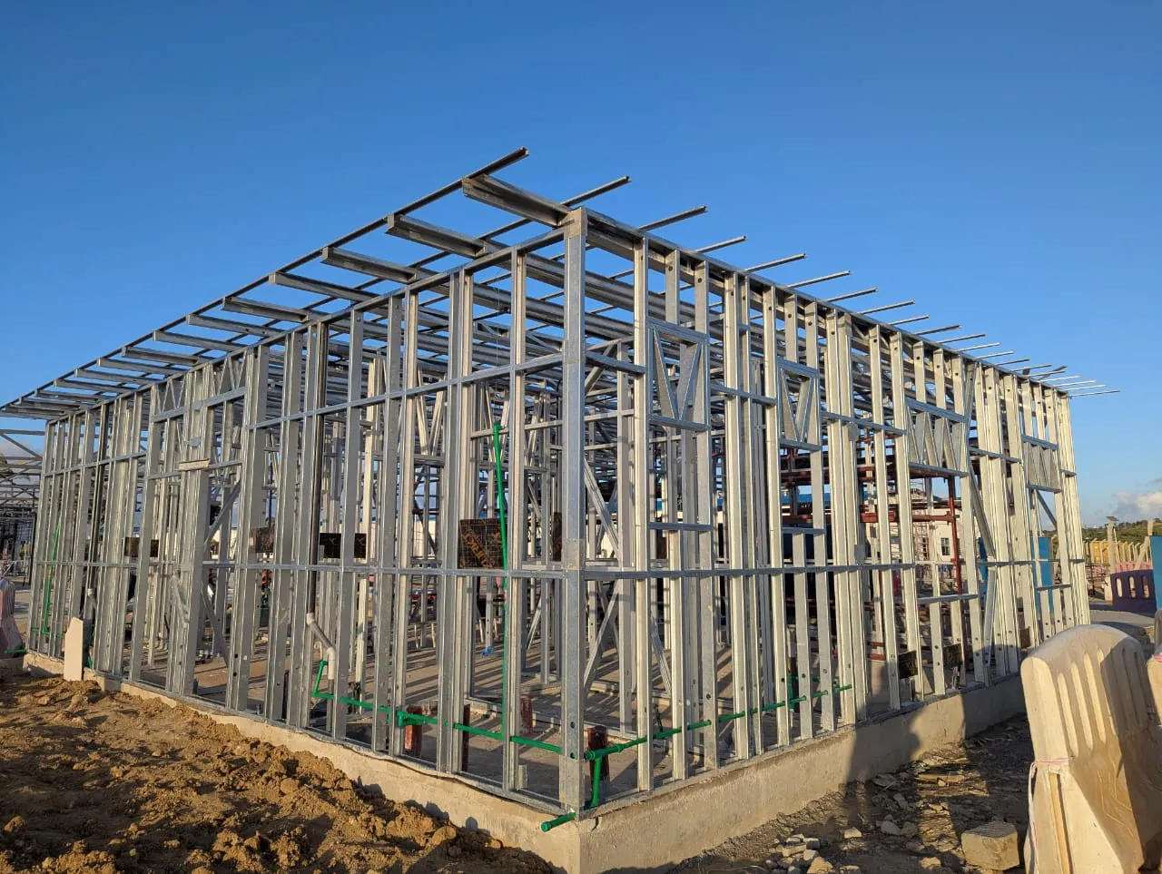 A complete light gauge steel frame standing on a concrete slab, walls braced and roof members in place, with green service pipes stubbed up through the floor
